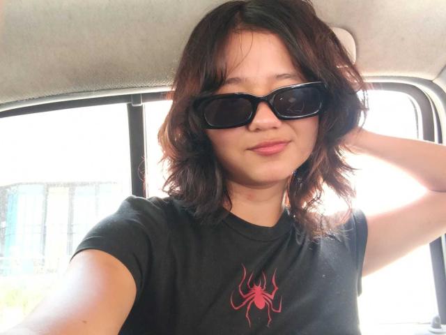 Sophie Smith, avec ses cheveux noirs, souriante, portant un t-shirt gris à motif, un pantalon noir et un sac à dos, se tient sur un pont en bois. Ses mains sont dans ses poches. Un arrière-plan de forêt..