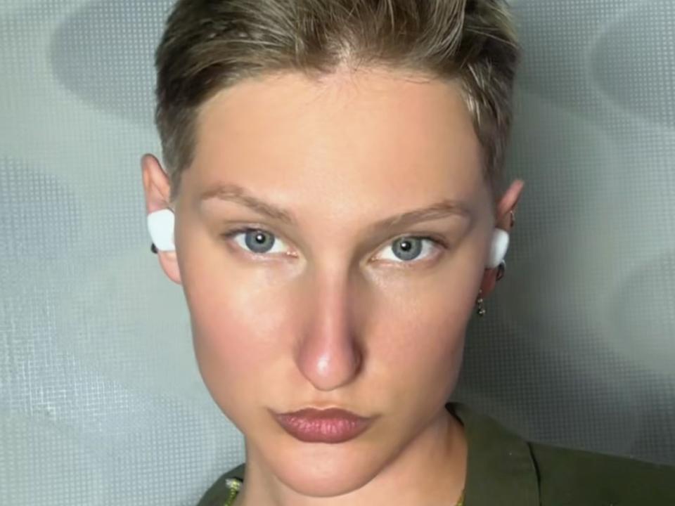 EmaJames con un reggiseno nero, che si sporge in avanti, mostrando il seno, capelli scuri, rossetto rosso e un sorriso malizioso, con i suoi occhi marroni rivolti verso l'alto. Il suo sedere è leggermente visibile..