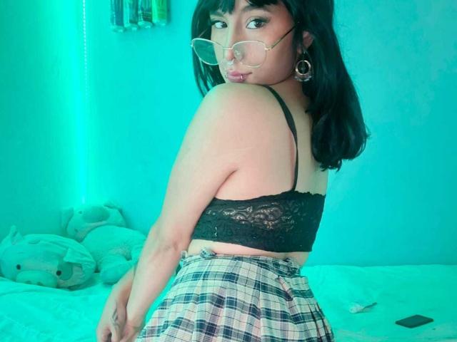 SerineHotAss with black lace bra, tattoos on chest and neck, winking, tongue out, glasses, nose ring, and earrings, hand in hair, greenish lighting.