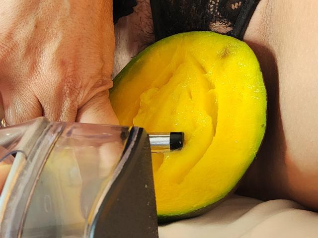 La mano de ScarletthVega sostiene una rodaja de mango amarillo con un palillo de metal, junto a un recipiente de plástico transparente que muestra su trasero cubierto con lencería negra de encaje..