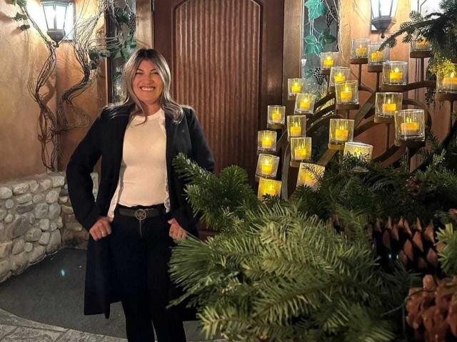 LunaBeya stands smiling with hands on hips, wearing a black coat, white shirt, and black pants, surrounded by lit candles and pine branches, in a warmly lit room.