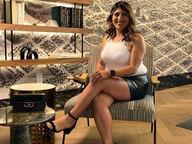 LunaBeya, with medium tits in a white tank top and shiny black skirt, sits on a chair, smiling, legs crossed, wearing black heels, in a stylish room.
