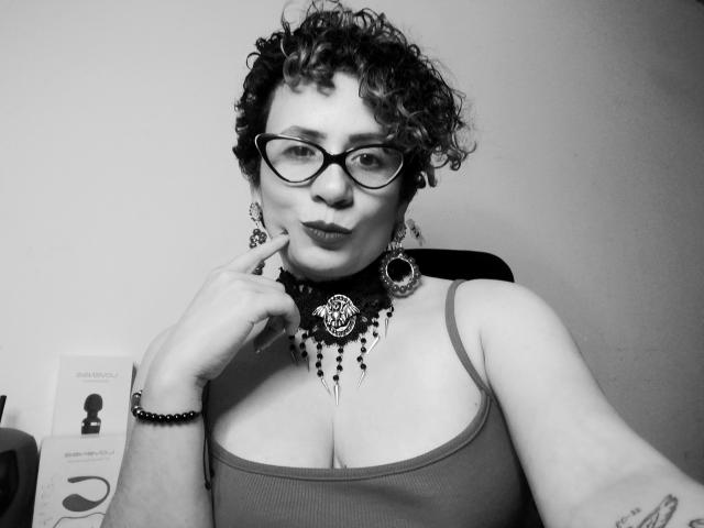Fotografía en blanco y negro de DaneshkaNaughty69 con cabello rizado, gafas, collar y pulsera, haciendo pucheros y tocándose la mejilla con un dedo, vistiendo una camiseta sin mangas, mostrando sus pechos..