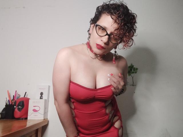 DaneshkaNaughty69, avec des cheveux bruns bouclés, des lunettes, une robe rouge, montrant ses seins, tenant un objet, des ongles roses, un collier, des boucles d'oreilles, et un regard séduisant. Une table avec un téléphone rouge et une boîte blanche..