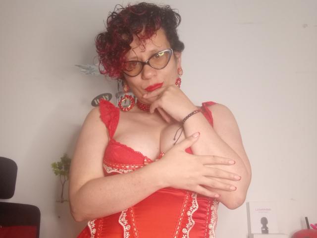 DaneshkaNaughty69 with curly red-black hair, glasses, red lace corset, red earrings, and red lipstick, holding her tits, hand on her chin, in a slightly provocative pose.