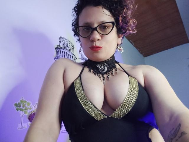DaneshkaNaughty69 with curly pink-tipped hair, glasses, red lipstick, black halter dress with gold trim, showing large tits, black lace choker, tattoo on right arm, serious expression.