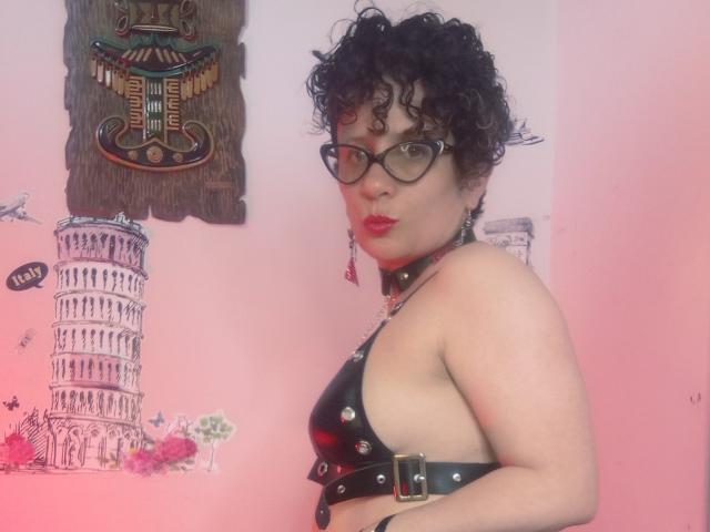 DaneshkaNaughty69, avec ses cheveux noirs bouclés, ses lunettes, son rouge à lèvres rouge, son harnais en cuir noir avec des clous métalliques et son collier noir, montrant sa poitrine nue et affichant un regard confiant et sexy..