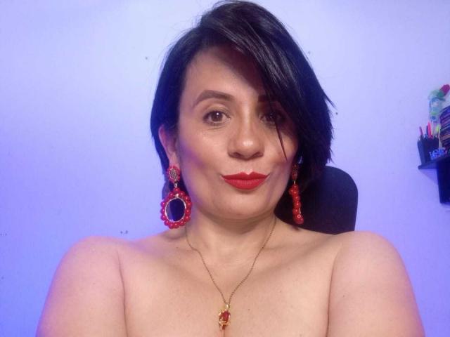 DaneshkaNaughty69 met kort zwart haar, rode oorbellen, rode lippenstift en een rode ketting, zonder top, waarbij haar blote schouders en een subtiele glimlach zichtbaar zijn. Haar borsten zijn gedeeltelijk zichtbaar..