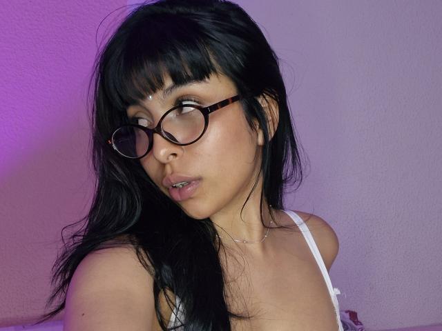 Ada Walton, com cabelos pretos longos, óculos e uma camiseta branca, mostrando seus seios, olhando para cima com uma luz rosa-roxo em seu rosto. Ela está usando um colar fino..