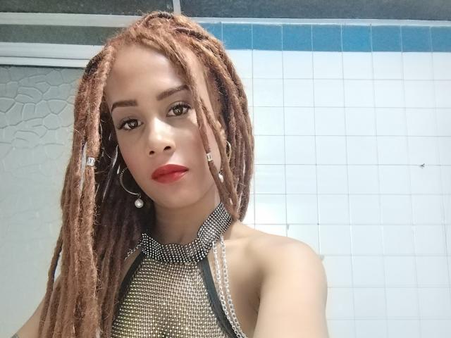 KendallQuinn with brown dreadlocks, red lipstick, hoop earrings, and a silver-studded halter top, showing her bare shoulders and upper chest. Her eyes are looking at the camera.