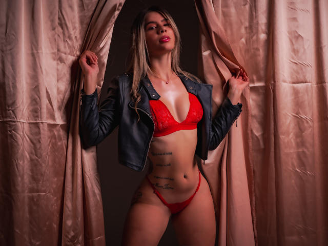 RileySwet stands in red lingerie with a black leather jacket, holding pink curtains. She's got tattoos on her side, and her tits are visible in the lace bra.