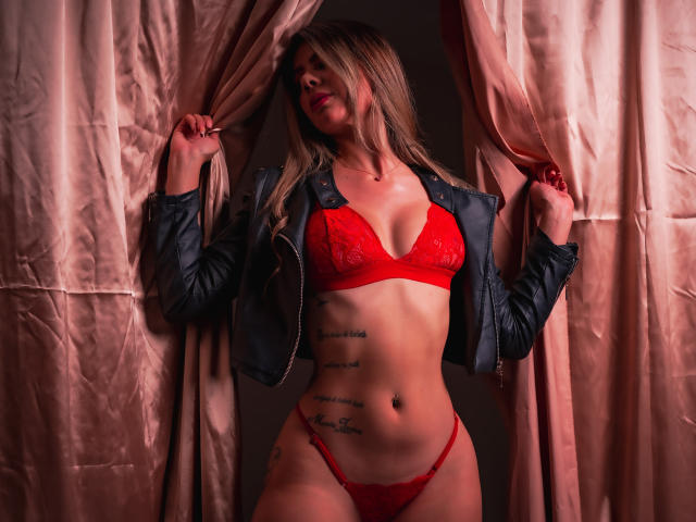 RileySwet vestindo um sutiã e calcinha de renda vermelha, jaqueta preta, segurando cortinas rosa, tatuagem no lado direito, cabelo loiro, batom vermelho, magra, seios pequenos, abdômen plano, sexy..