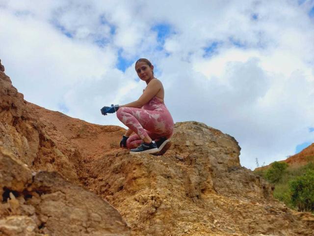 EliBaker crouches on rocky brown hill, wearing pink patterned pants, blue gloves, black shoes, smiling, with a cloudy blue sky behind. Her ass is visible.