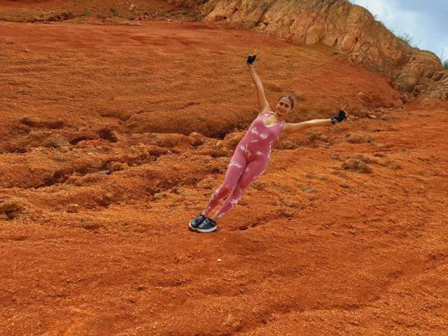 EliBaker is standing on red rocky ground, wearing pink workout pants and a tank top, with arms raised and black gloves, in a rocky, orange landscape.