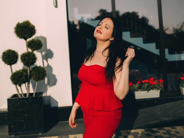 CassieRose met lang donker haar, gekleed in zwarte, open lingerie en een choker, zittend op een houten vloer, met gekruiste benen, lachend, waarbij haar borsten en vagina zichtbaar zijn, in warm licht..