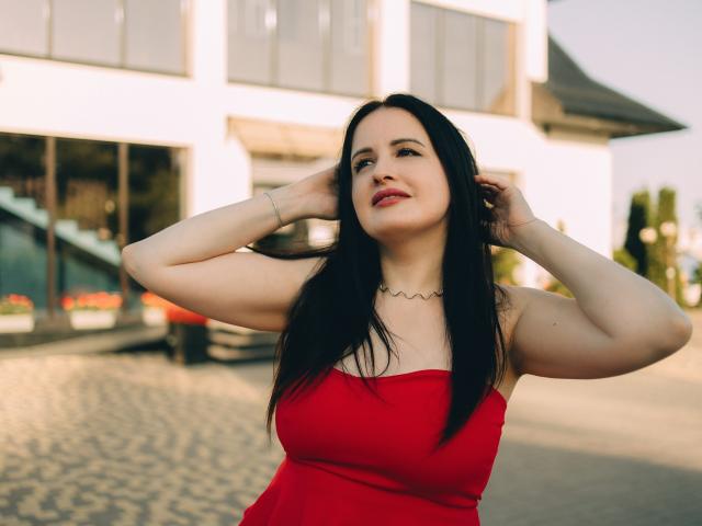 TayaBoret mit langem, schwarzem Haar, rotem Kleid und gerafftem Oberteil, die sich die Haare hält, die Augen geschlossen hat und lächelt, steht vor einem schwarzen Pflanzgefäß mit grünen Büschen, im Sonnenlicht, mit roten Blumen..