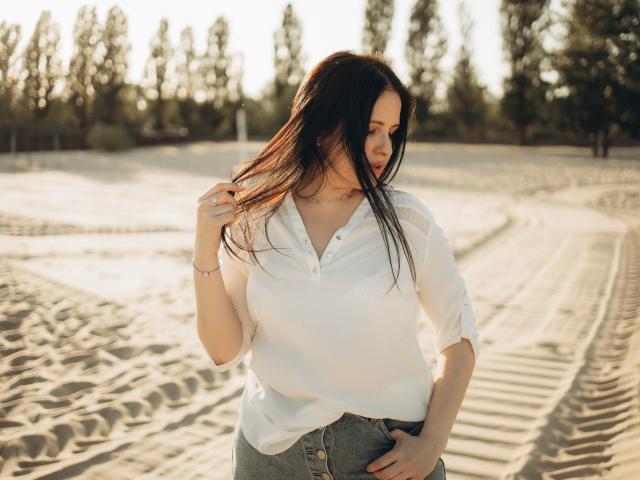 TayaBoret with long dark hair, white blouse, and buttoned denim skirt, hand in hair, standing in sunlit, sandy area, looking to the side, soft golden light.
