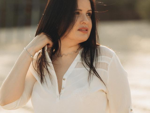 TayaBoret with long black hair, white blouse, and gray pants, holding her chest, sunlight on her face, slight smile, right hand up, left hand on blouse, relaxed.