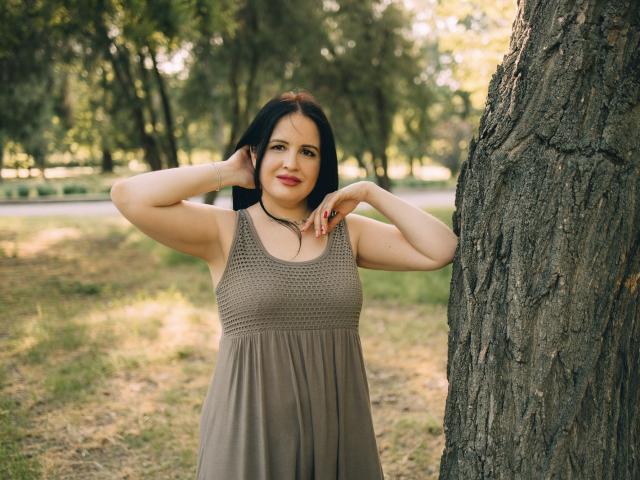 TayaBoret, mit langem, schwarzem Haar und mittlerer Brustgröße, steht in einem braunen Kleid vor einem Baum, wobei Sonnenlicht durch die Blätter scheint. Sie hat ein sanftes Lächeln im Gesicht und den rechten Arm angehoben..
