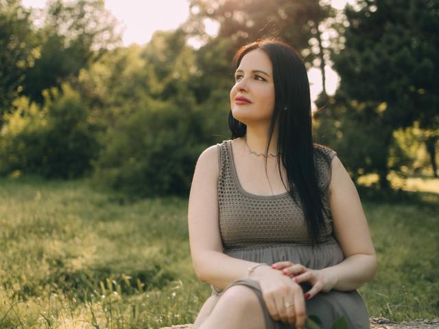 TayaBoret se tient contre un arbre, les bras levés, vêtue d'une robe marron sans manches, avec un collier noir et du rouge à lèvres, elle a une poitrine de taille moyenne et des cheveux foncés, dans un parc ensoleillé..