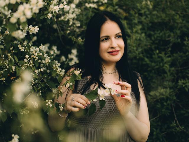TayaBoret, avec ses cheveux noirs, son rouge à lèvres rouge et son sourire charmant, tient des fleurs blanches. Elle porte un haut en dentelle et un collier ondulé, avec un feuillage vert en arrière-plan..