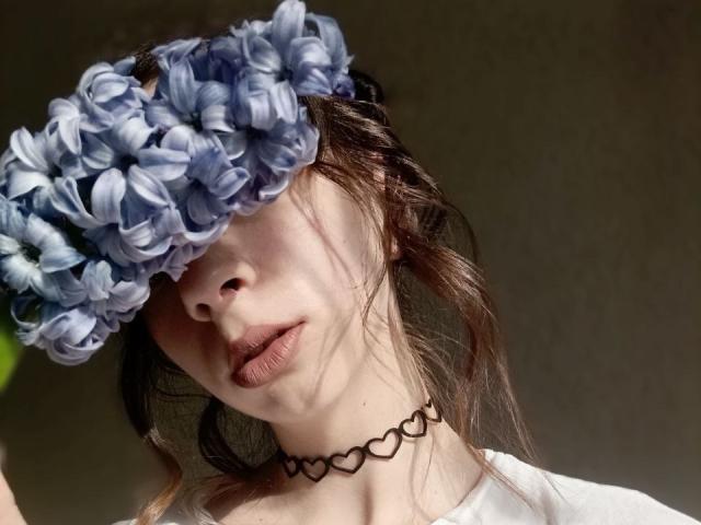 WollyMolly met een blauwe bloemenhoofdband die haar ogen bedekt, een zwarte hartketting en een witte blouse, waarbij haar volle lippen en licht golvende bruine haar zichtbaar zijn..