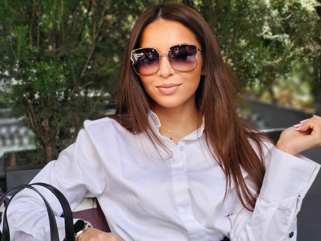 KayaOcean with long brown hair, wearing a white button-up shirt and dark sunglasses, smiling slightly with her hand near her face, sitting outdoors. Her tits are small.