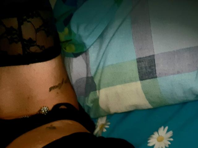 De onderbuik van MaliKanal, met een tatoeage en een zwarte kanten top, met een zwarte bh en een buikpiercing, liggend op een bed met een blauw-groen geruit patroon..