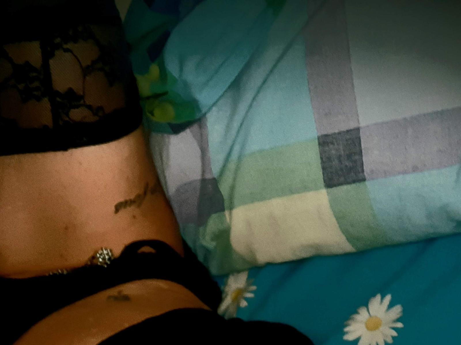 A parte inferior da barriga de MaliKanal, mostrando uma tatuagem e uma blusa de renda preta, com um sutiã preto e um piercing no umbigo, deitada em uma cama com padrão quadriculado azul e verde..
