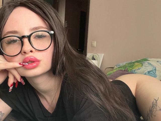 EllaLaLarsen, avec des lunettes, du rouge à lèvres rose et des ongles rouges, faisant la moue, portant un haut noir, avec un tatouage sur le bras droit, allongée sur un lit, les fesses en l'air, cheveux bruns, peau pâle, chambre à coucher..