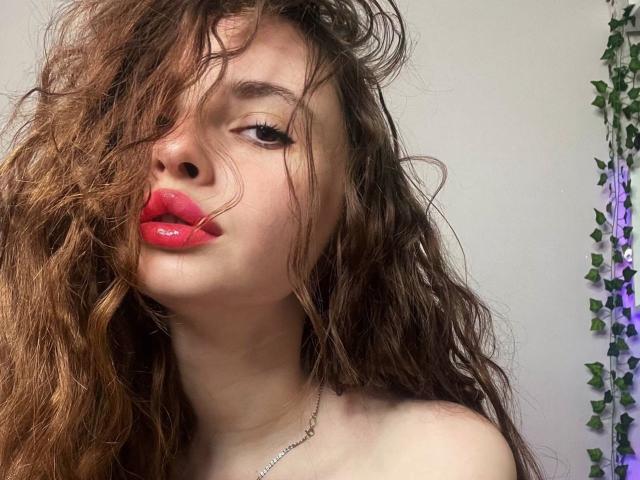 EllaLaLarsen with messy brown hair, red glossy lips, and a silver necklace, looking slightly to the side. Her bare shoulders are visible.