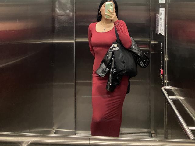 Elizaa, con un vestito rosso aderente, tiene in mano una giacca nera, è in piedi in un ascensore e sta scattando un selfie con un telefono verde, mostrando il sedere e i seni..