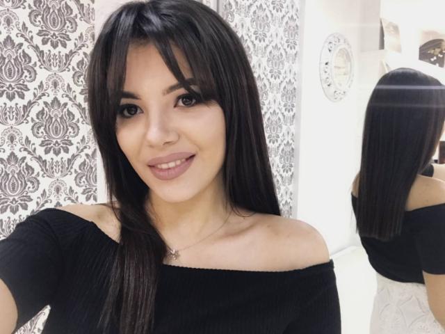 OnlyXandra, con i suoi lunghi capelli neri, la frangia e un sorriso, indossa una maglietta nera con una spalla scoperta, i suoi seni appena coperti, in un selfie allo specchio, che mostra la sua schiena nel riflesso..