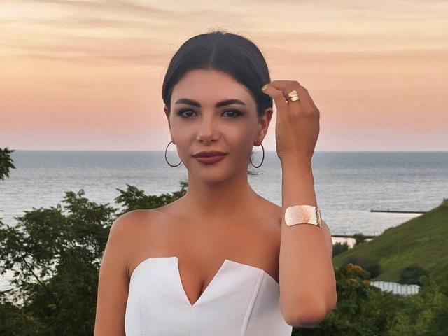 OnlyXandra, con i capelli scuri, orecchini a cerchio, top bianco senza spalline, braccialetto dorato e anello, si tiene i capelli, mostrando il seno, con uno sfondo di oceano al tramonto..