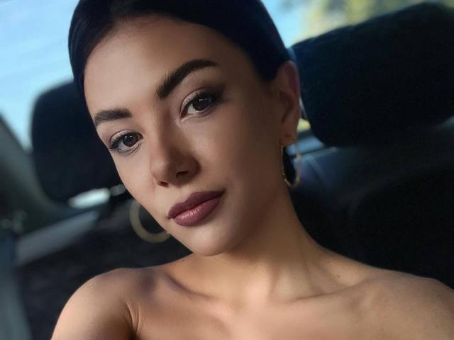 OnlyXandra, mit dunklem Haar, Reifenohrringen und unbedeckten Schultern, hat glatte Haut, dunkle Augen und rosafarbenen Lippenstift und blickt direkt in die Kamera mit einem leichten Lächeln..