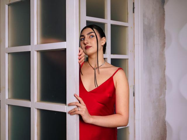 MirrorOfHeartI con cabello oscuro, vestido rojo y collar, de pie junto a una puerta de vidrio con marco blanco, sosteniéndola con una mano, mirando hacia un lado con una ligera sonrisa..