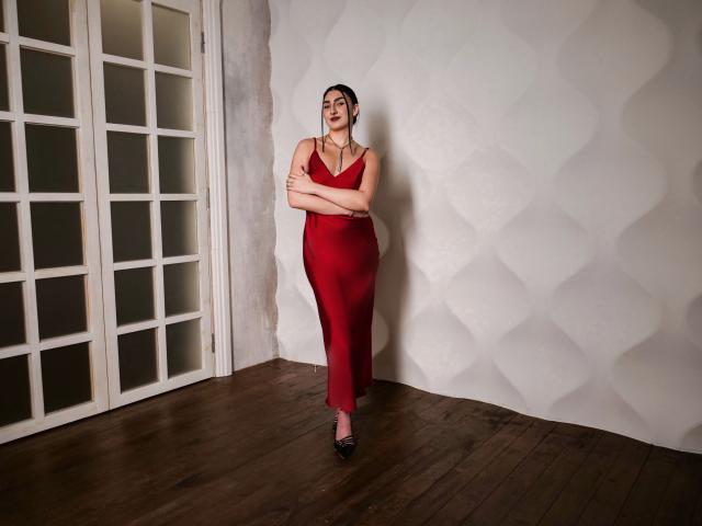 MirrorOfHeartI está de pie, vestida con un vestido rojo, con los brazos cruzados, llevando tacones negros y una diadema negra, luciendo segura de sí misma, en una habitación con suelos de madera y paredes blancas con forma de onda..