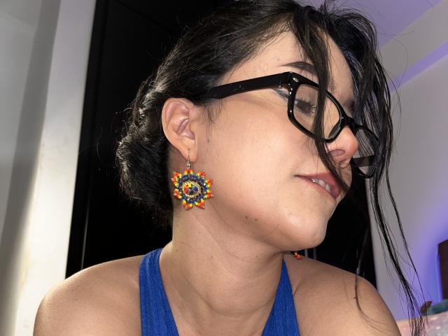 DeisyWest with dark hair, glasses, and blue top, showing off colorful, round earrings, looking to the side with a slight, relaxed smile. Her face has a small mole.