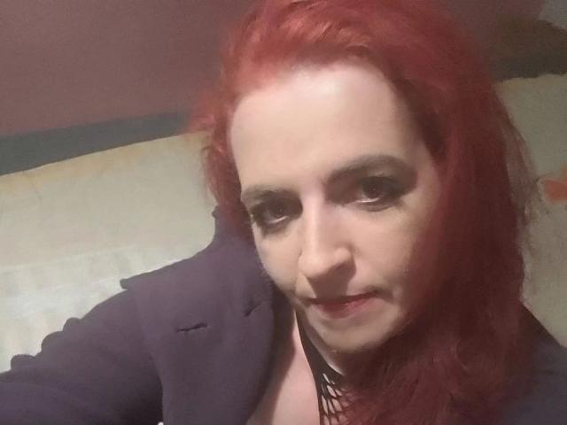 IsaWild, con i capelli rossi, indossa un blazer scuro, ha la pelle chiara e un trucco scuro per gli occhi, guarda la fotocamera con un leggero sorriso, mostrando un piccolo piercing al naso..
