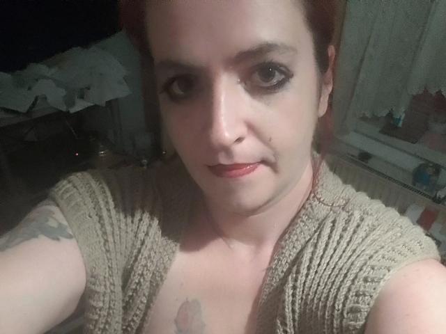 IsaWild, con cabello rojo, maquillaje oscuro y labial rojo, vestida con una blusa beige de punto, mostrando tatuajes en su pecho y brazo, en una habitación con poca luz..