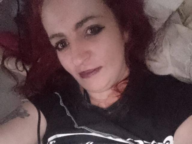 IsaWild-hot tiene el pelo rojo y rizado, delineador de ojos oscuro y lápiz labial morado, está acostada con una camiseta negra, mostrando un tatuaje en su brazo izquierdo, y mirando a la cámara..