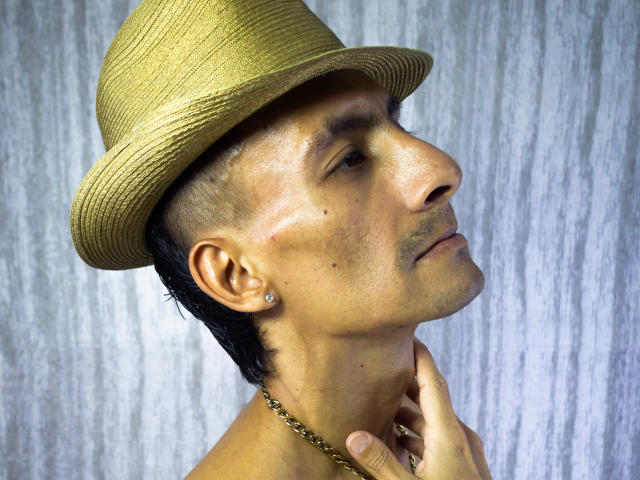 TheRoomSex69 ist ein Mann mit gebräunter Haut, der einen Strohhut mit Goldverzierungen, kurze schwarze Haare, Ohrringe und eine goldene Kette trägt. Er blickt nach oben und hat eine Hand in der Nähe seines Halses..