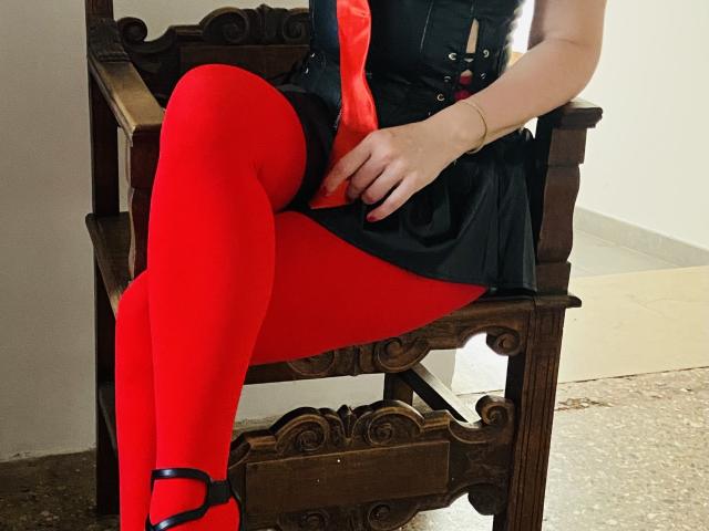 O traseiro de LexyMoon, coberto por uma calcinha vermelha, está sentado em uma cadeira de madeira, usando sapatos pretos e uma gravata vermelha, vestindo um vestido preto e meias vermelhas. Sua mão está apoiada em sua coxa..