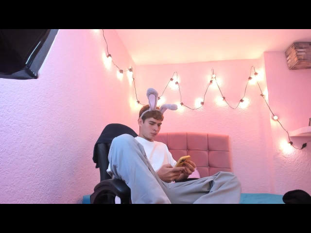 AngelHub, con orejas de conejo, vistiendo una camisa blanca y pantalones grises, está sentado en una silla comiendo un sándwich, mirando hacia abajo, en una habitación con paredes rosas y luces de cadena..
