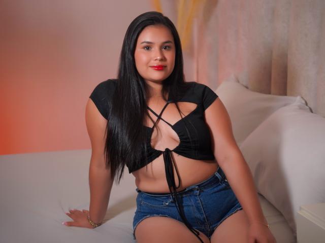 AngelinaBlanche with red lipstick, wearing a white, cherry-patterned, sheer top, showing her small tits and a gold ring on her right hand, in a kitchen, with one arm up.