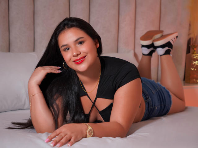 AmilyJones with long black hair, red lipstick, black crop top showing her tits, and denim shorts, sitting on a bed, looking at the camera with a slight smile.