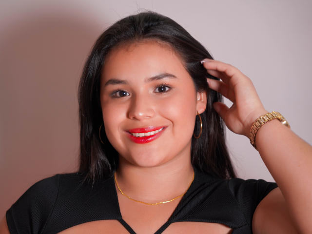 Amily Jones, con cabello negro largo, lápiz labial rojo y una blusa negra, está acostada en una cama, mostrando su trasero, con pantalones cortos vaqueros azules y tacones negros y blancos, y sonriendo a la cámara..