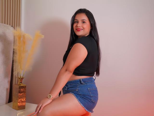 AmilyJones has medium brown skin, black hair, red lipstick, and a gold necklace. She's smiling with her hand in her hair, wearing a black top and gold bracelet.