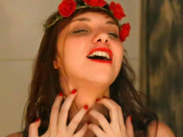 Amily Jones, con lunghi capelli neri, rossetto rosso, reggiseno verde, che si tiene i seni, collana d'oro, unghie rosa, braccialetto, sorridente, in interni. Seni, reggiseno verde, labbra rosse, gioielli..