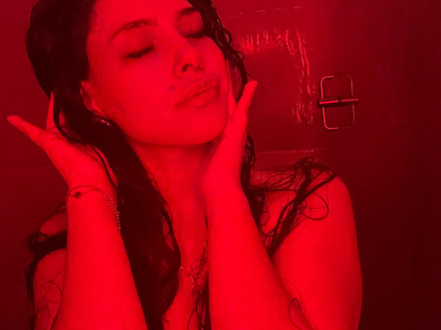 LorreineMontclair with a pink towel on her head, white towel wrapped around her ass, shaving her armpit with a pink razor in a gray-tiled bathroom.